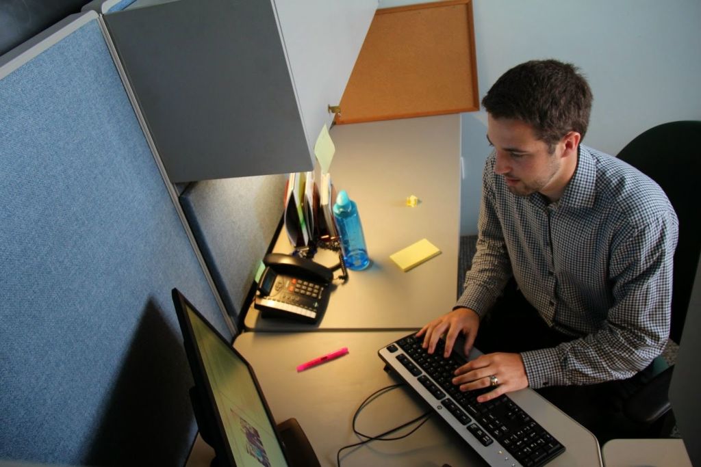 Hombre joven escribiendo en el teclado de su computadora dentro de un cubiculo de oficina