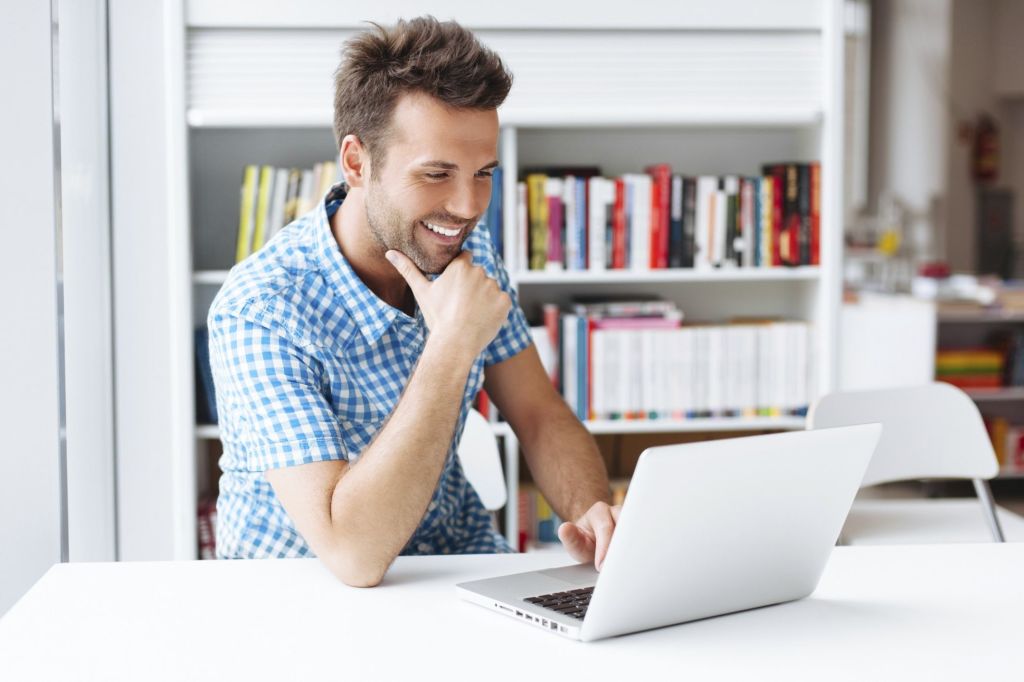 Hombre joven sentado a su mesa de oficina con la barbilla apoyada en su mano mientras mira su laptop sonriente