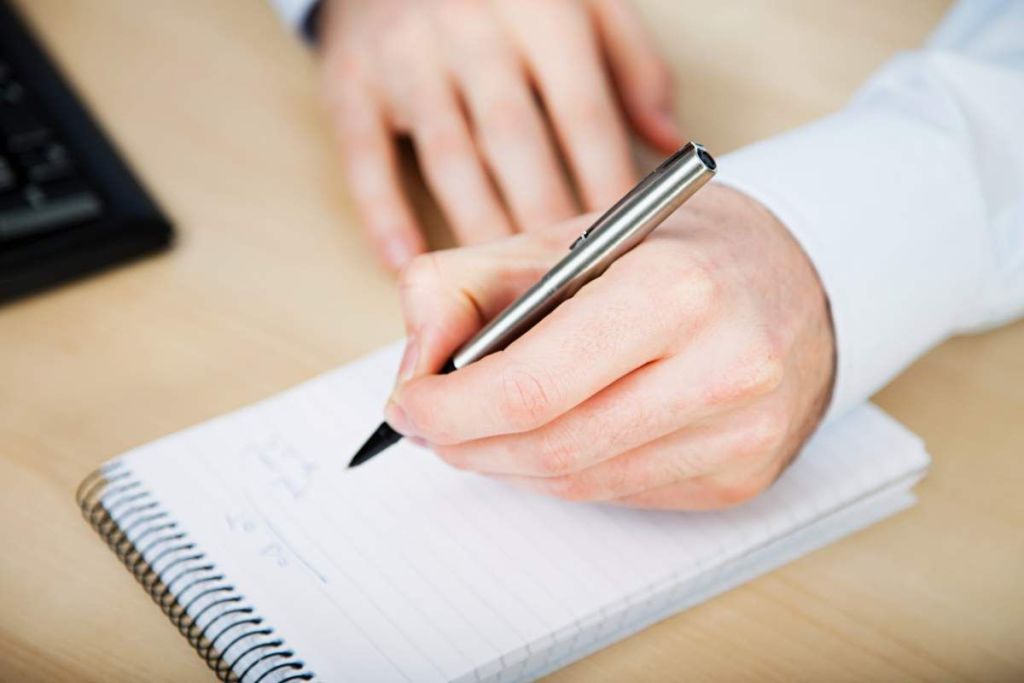 Hombre sosteniendo en su mano una pluma de escribir y escribiendo sobre un bloc de notas