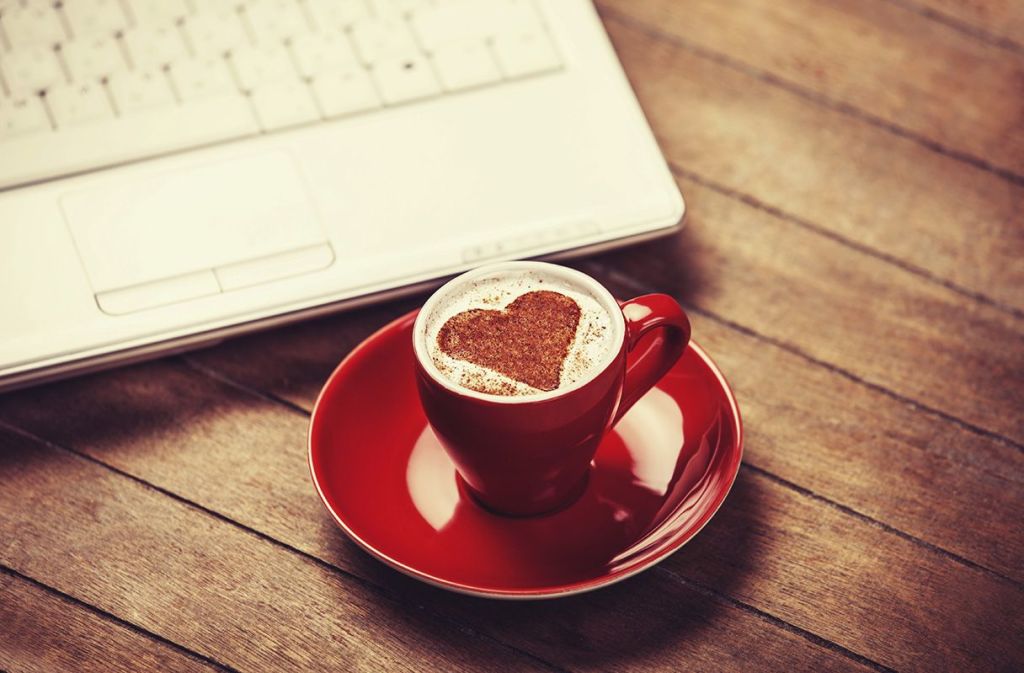 Una taza roja sobre un plato rojo con cafe dentro y la espuma del cafe tiene formado un corazon rojo encima de una mesa al lado de un portatil