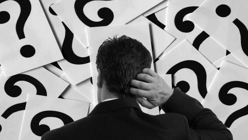 Hombre en blanco y negro rascandose la cabeza mientras mira a una pared llena de interrogaciones negras