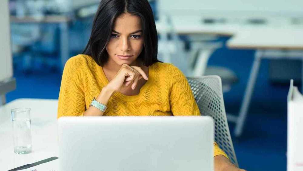 Mujer observando su computadora con la mano en su barbilla.
